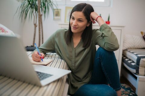 Young woman writing notes and paying utility bills on laptop